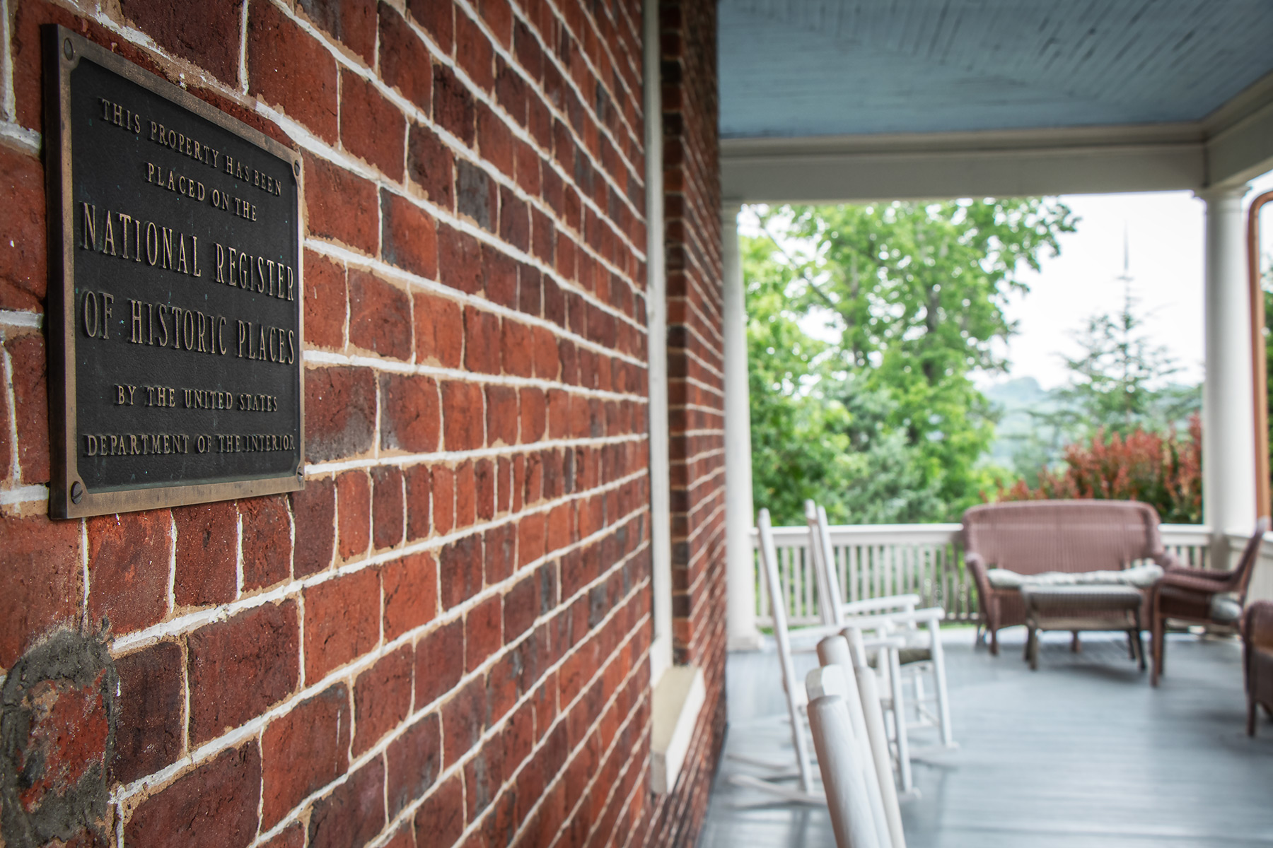 The National Register of Historic Places bronze plaque mounted on the original 1847 brick wall of The Reynolds Mansion, with the wraparound porch and rocking chairs visible in the background