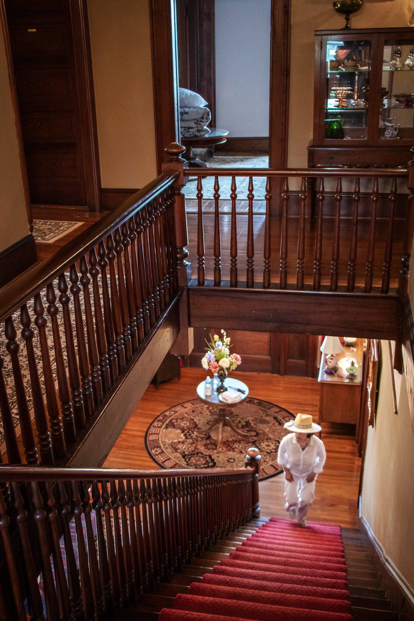View looking down the Colonial Revival staircase of The Reynolds Mansion — a guest ascending the crimson-carpeted stairs, turned balusters framing the entrance hall below with its Persian rug and fresh flowers