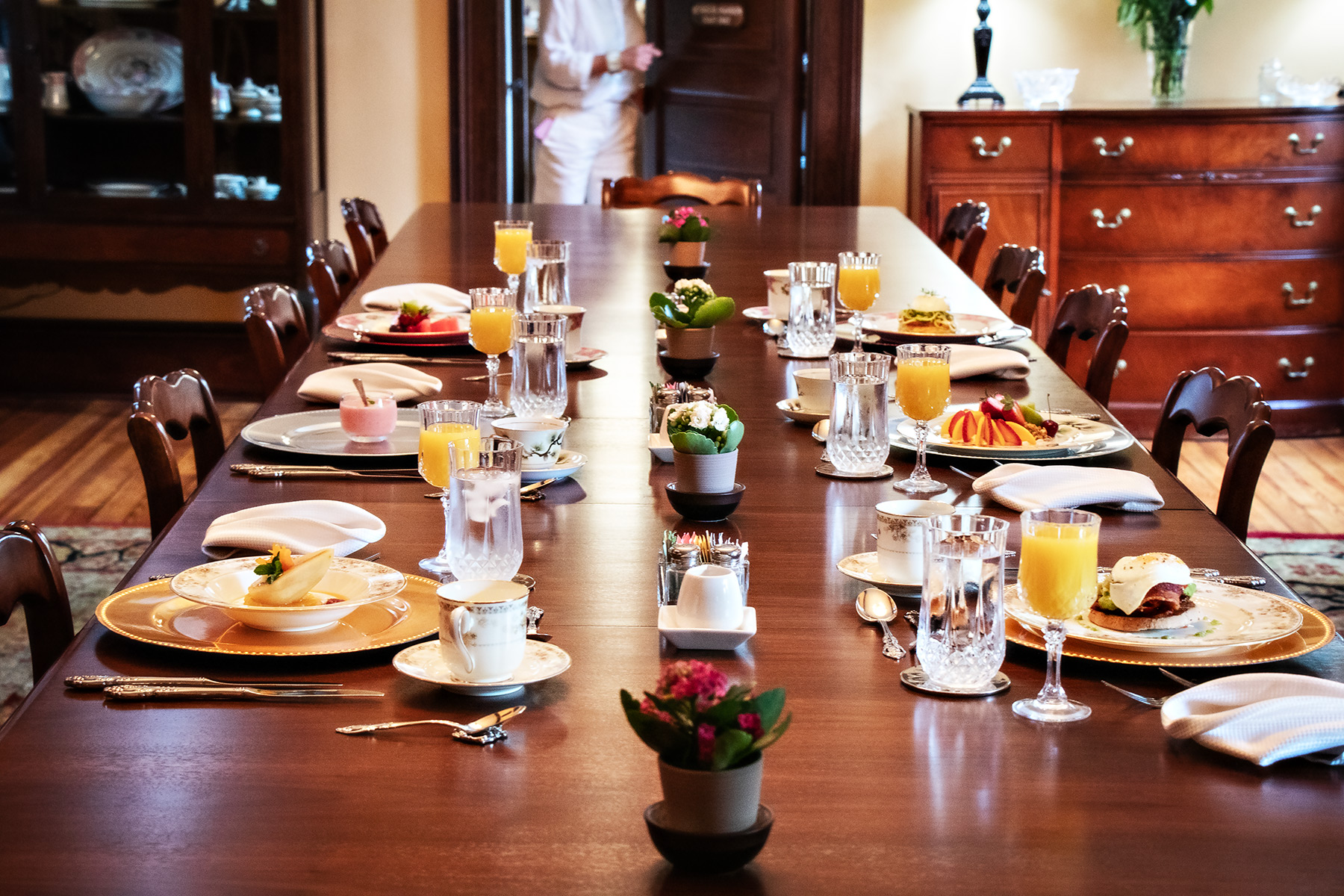 Breakfast service at The Reynolds Mansion — the mahogany table set with plated courses, crystal stemware, and fresh flowers as Chef Paul enters the grand dining room