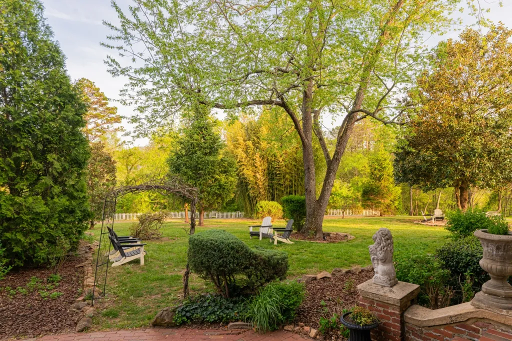 A quiet corner of the back garden at golden hour, with a swing, Adirondack chairs, a stone lion, and a bamboo grove beyond a white picket fence.