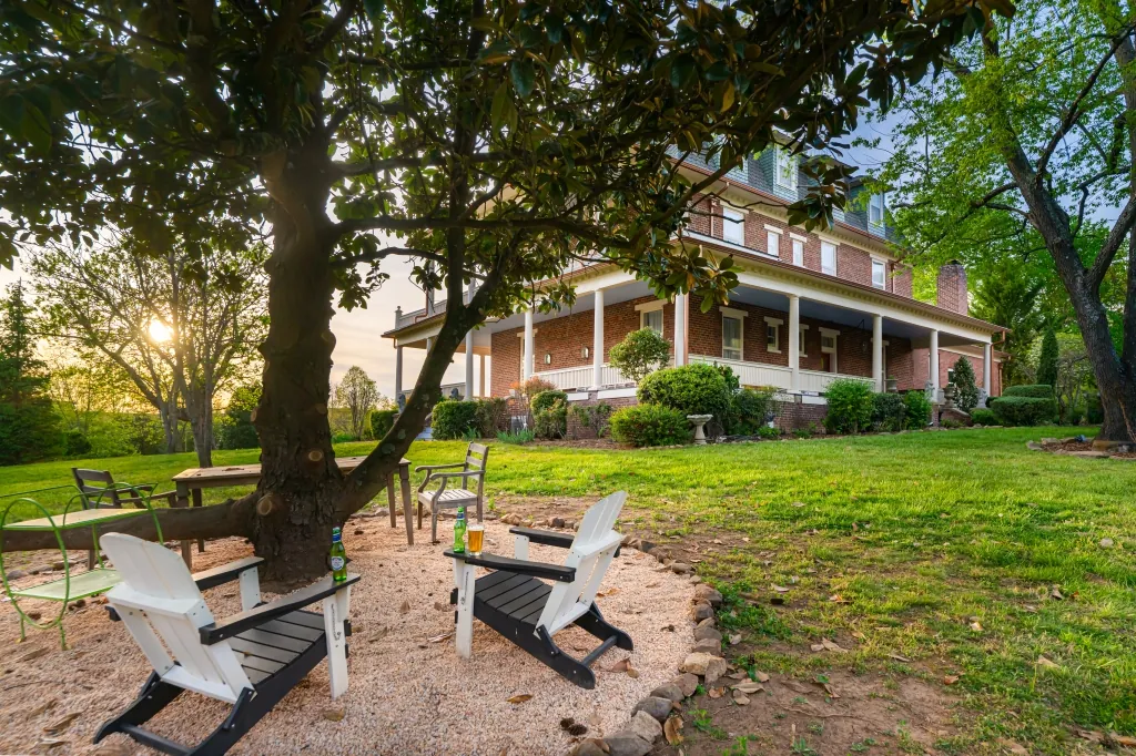 White and dark Adirondack chairs gathered under a spreading magnolia tree at sunset, with the mansion&rsquo;s porch behind.