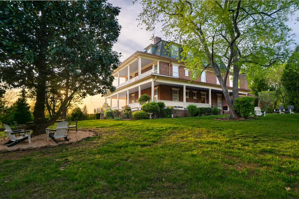 The brick mansion on a green rise at golden hour, framed by tall old trees, with Adirondack chairs set out on the lawn.