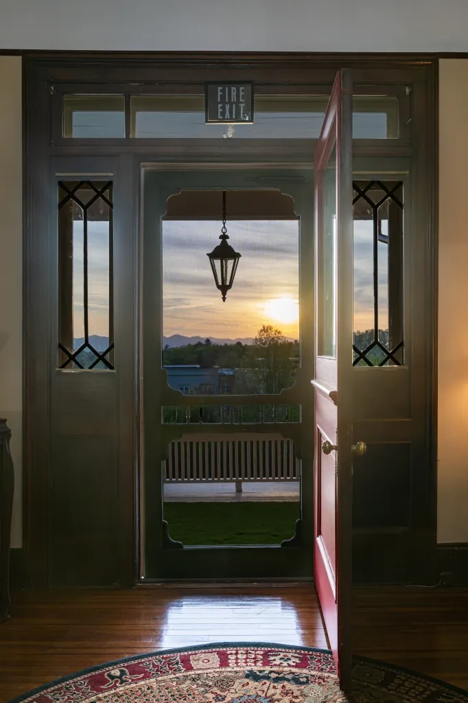 The open red front door of the mansion framing a sunset over the mountains, with leaded-glass sidelights and a polished wood floor.