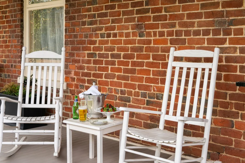 Two white rocking chairs on the porch beside a small table set with sparkling wine, a chilled beer, and fresh strawberries against the historic brick wall.
