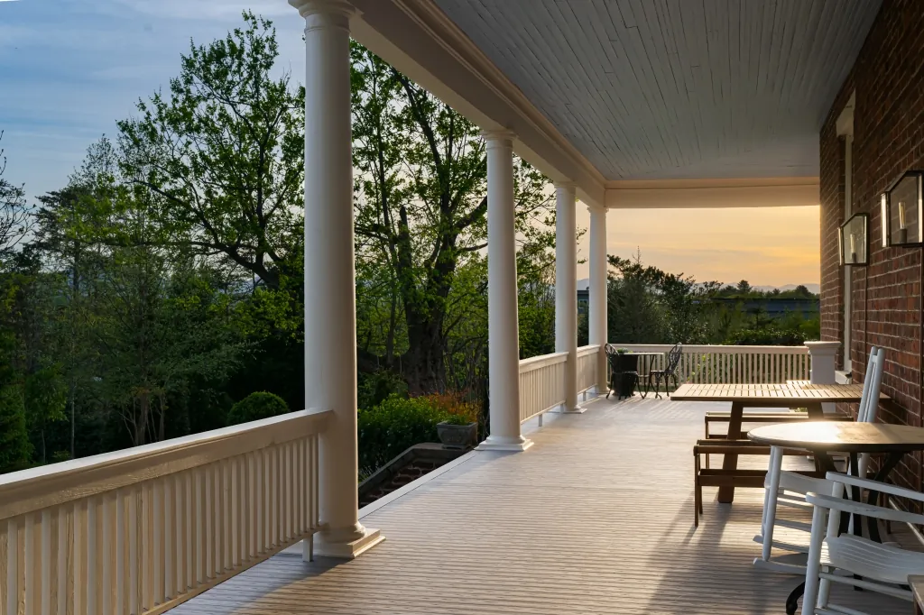 A teak table and chairs on the wrap-around porch in golden evening light, with a long view over the trees toward the mountains.