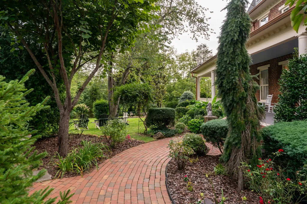 A curving brick path winding through the shaded summer garden, with a weeping evergreen and the Carriage House porch beyond.