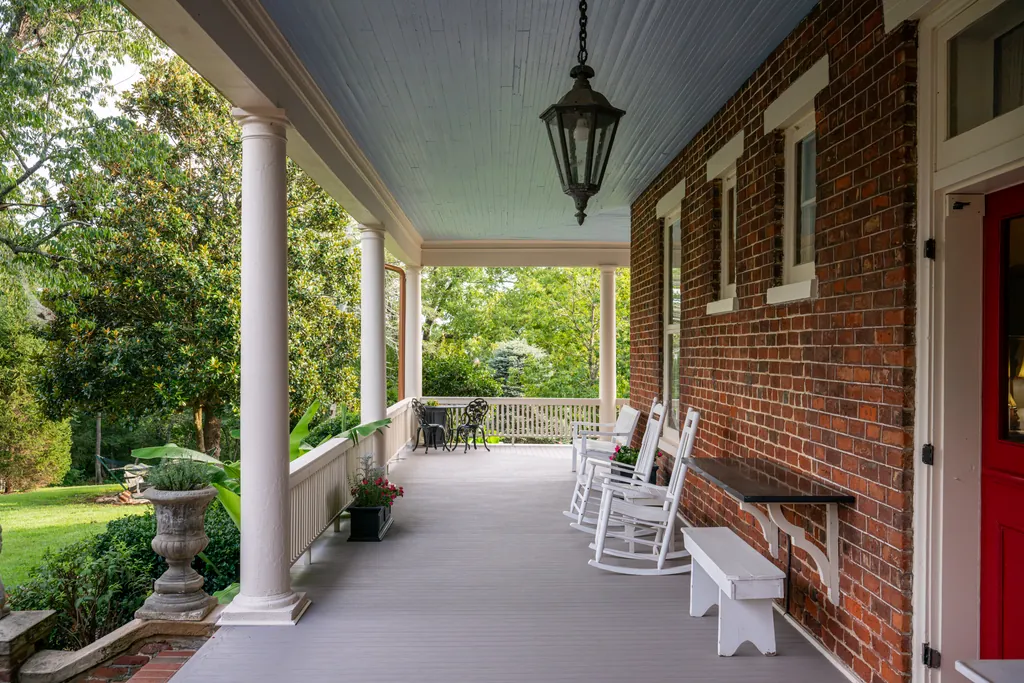 The columned front porch in summer, white rocking chairs and a bench set along the brick wall beside the red front door.