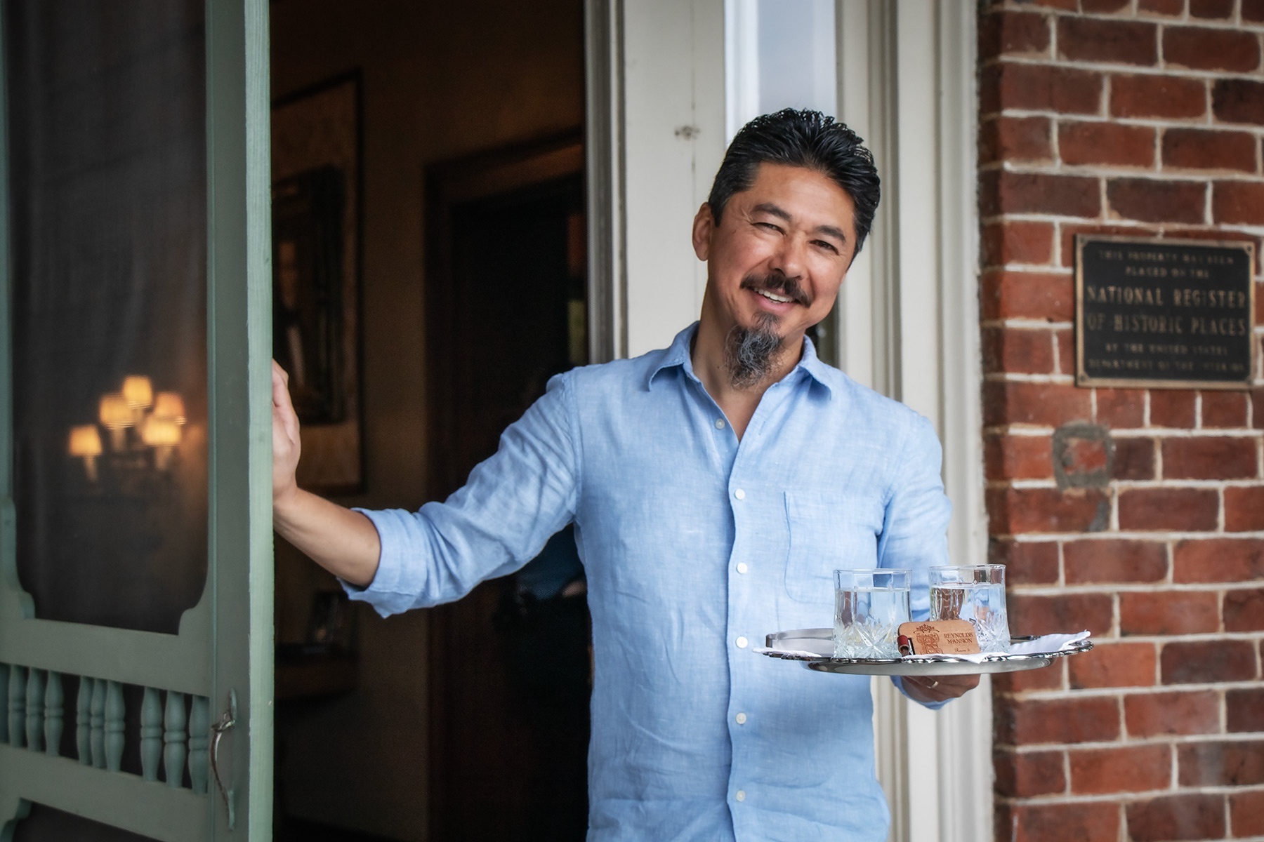 Paul Manshon welcoming guests at the front door of The Reynolds Mansion with a silver welcome tray — the National Register of Historic Places plaque visible on the 1847 brick wall