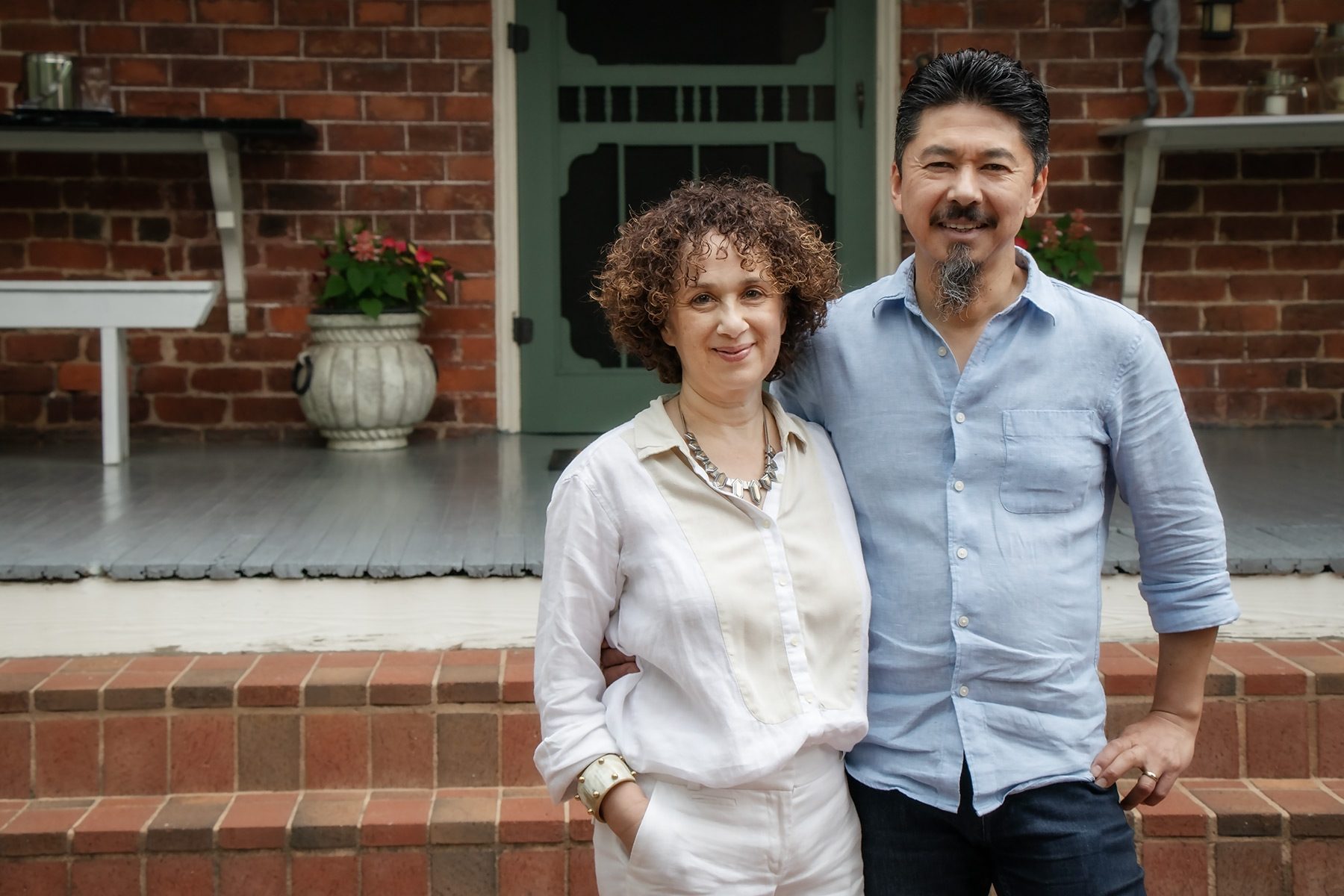 Paul and Mirlin Manshon standing together on the front porch of The Reynolds Mansion, the green screen door and 1847 brick wall behind them