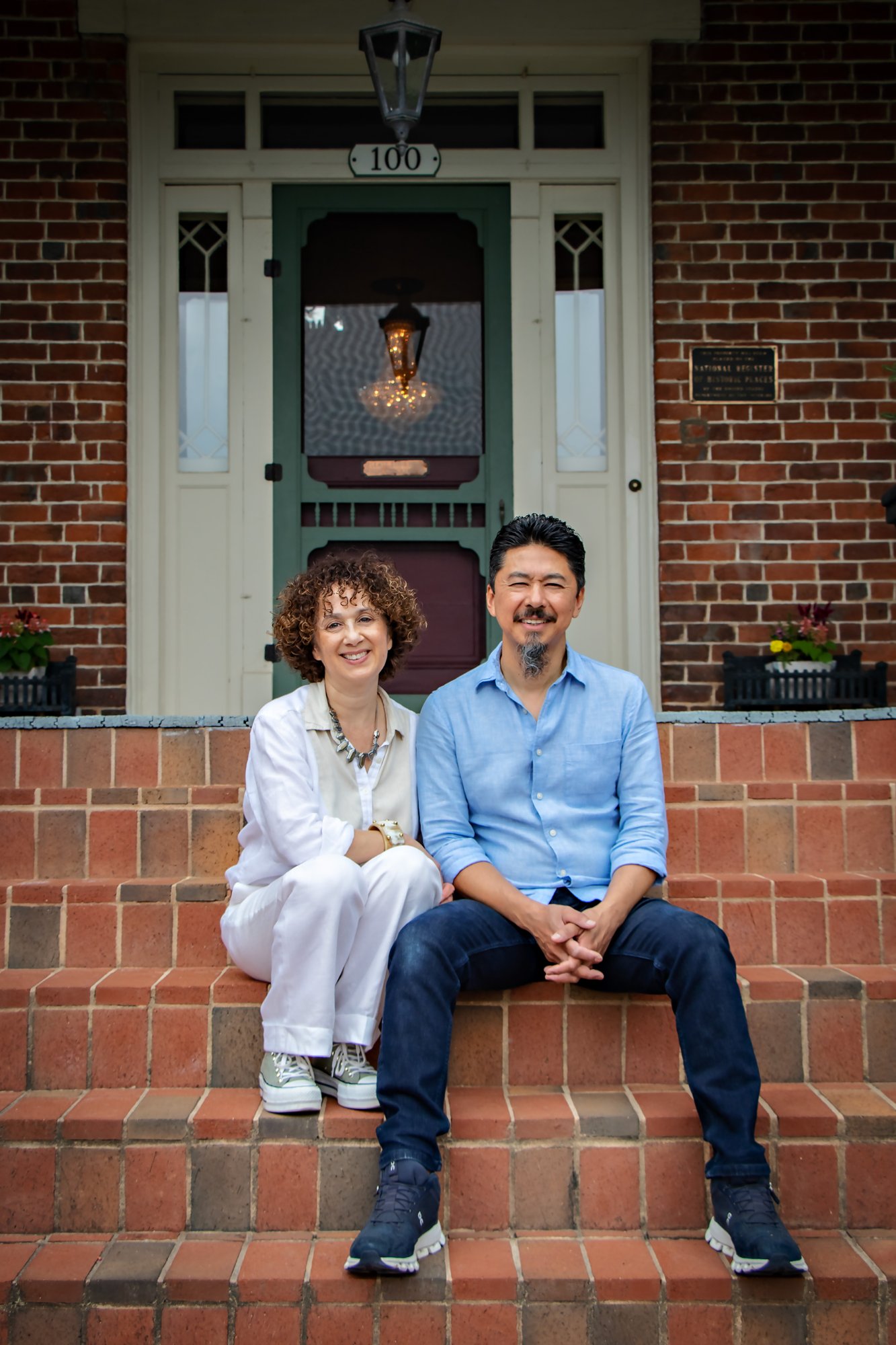 Paul and Mirlin Manshon sitting on the front steps of The Reynolds Mansion — the 1847 brick, front door, and address number 100 visible behind them