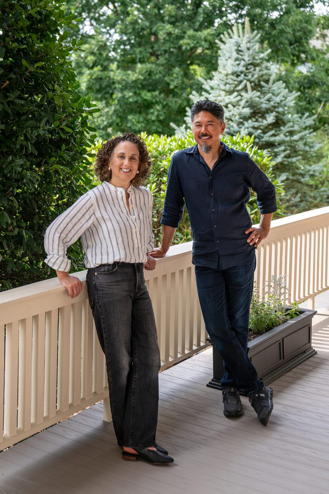 Paul and Mirlin Manshon on the wrap-around porch of The Reynolds Mansion, leaning on the white railing with the gardens behind them