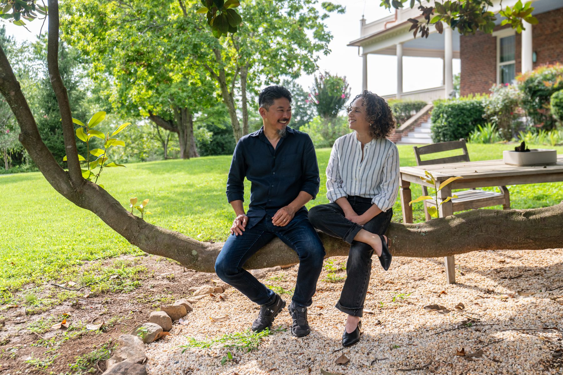 Paul and Mirlin Manshon, innkeepers of The Reynolds Mansion, sharing a laugh on a low tree limb in the garden with the mansion&rsquo;s porch behind them