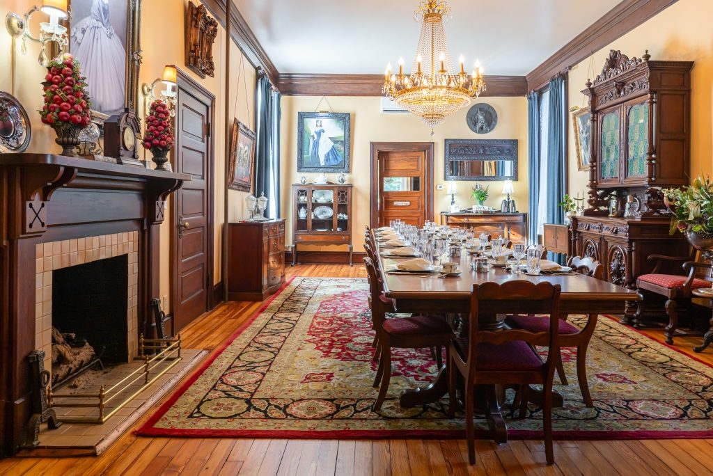 The grand dining room at The Reynolds Mansion — fireplace, crystal chandelier, antique china cabinet, and mahogany table set for breakfast