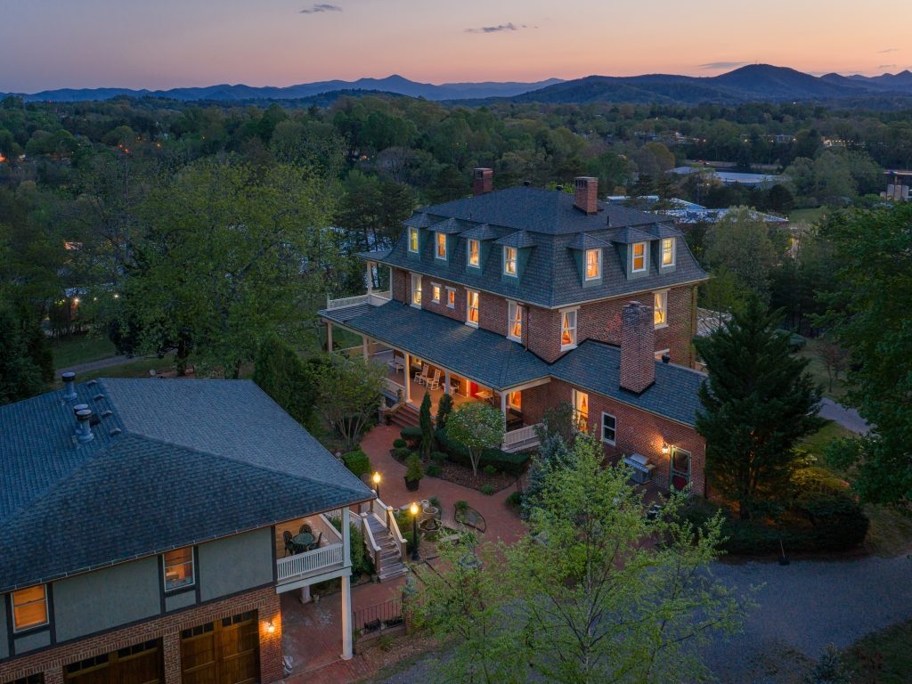 The Reynolds Mansion at twilight with warm light glowing from every window, set against the Blue Ridge Mountains and a rose-colored sunset sky