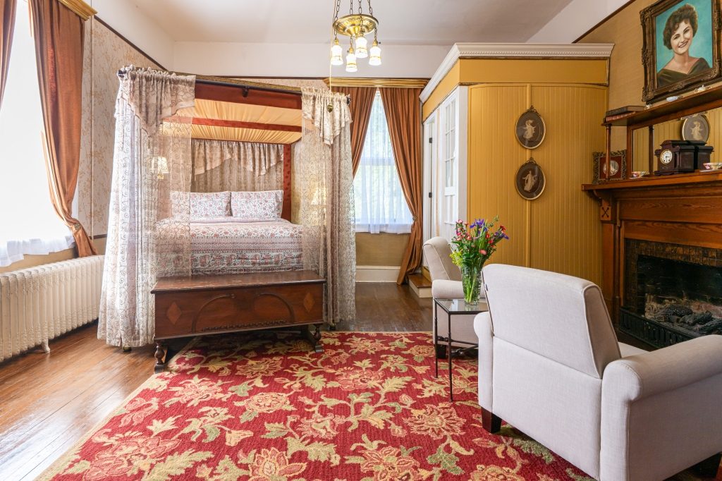 Glenda room — canopy bed with sheer curtains, golden wallpaper, period portrait, and gas fireplace at The Reynolds Mansion