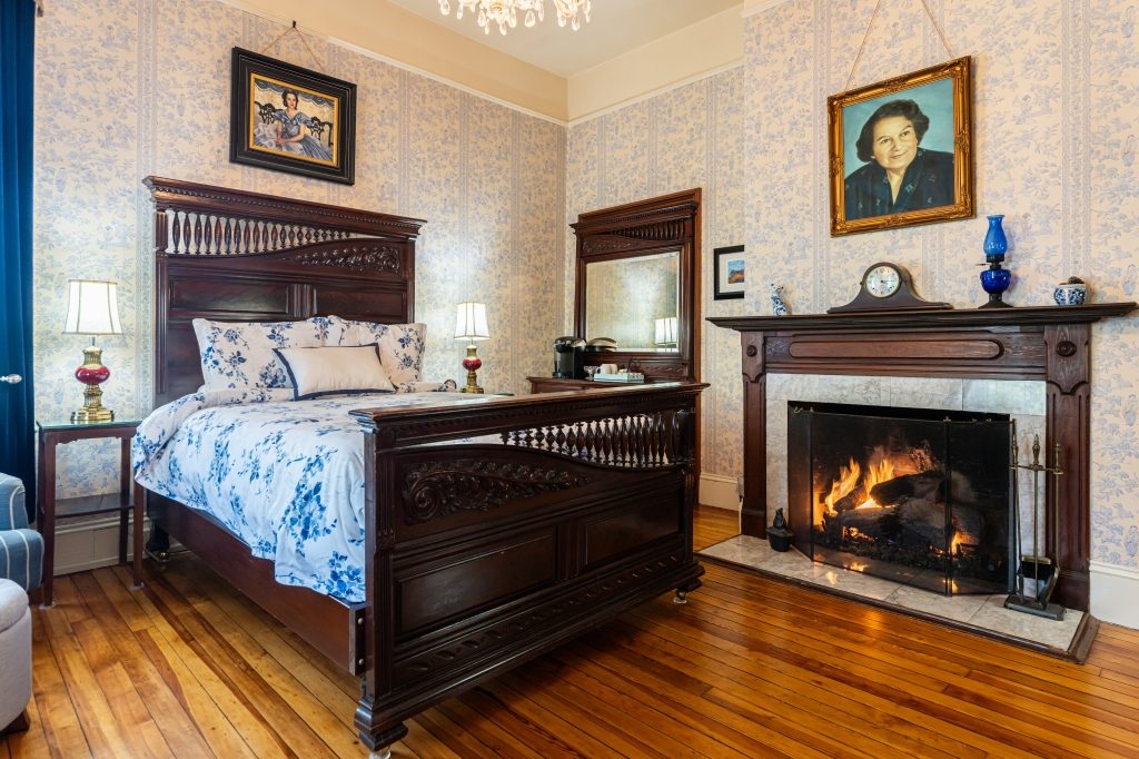 Lila room — antique queen bed against blue floral wallpaper with working gas fireplace and period portrait at The Reynolds Mansion