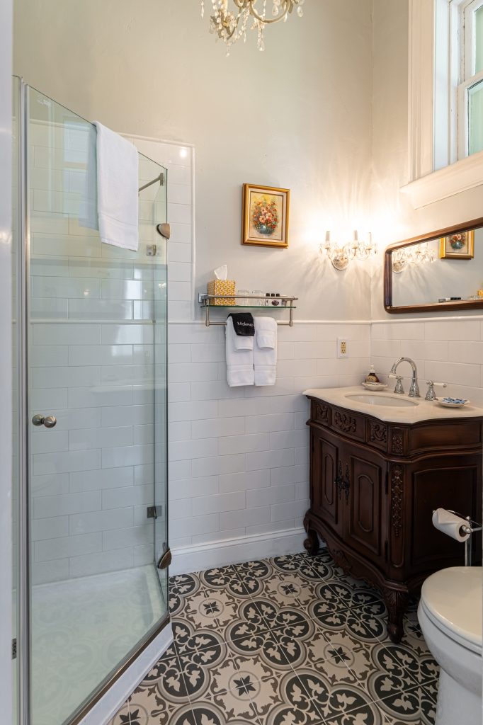Lila room private bathroom — glass shower stall with white subway tile, antique wood vanity with gold mirror, decorative tile floor at The Reynolds Mansion