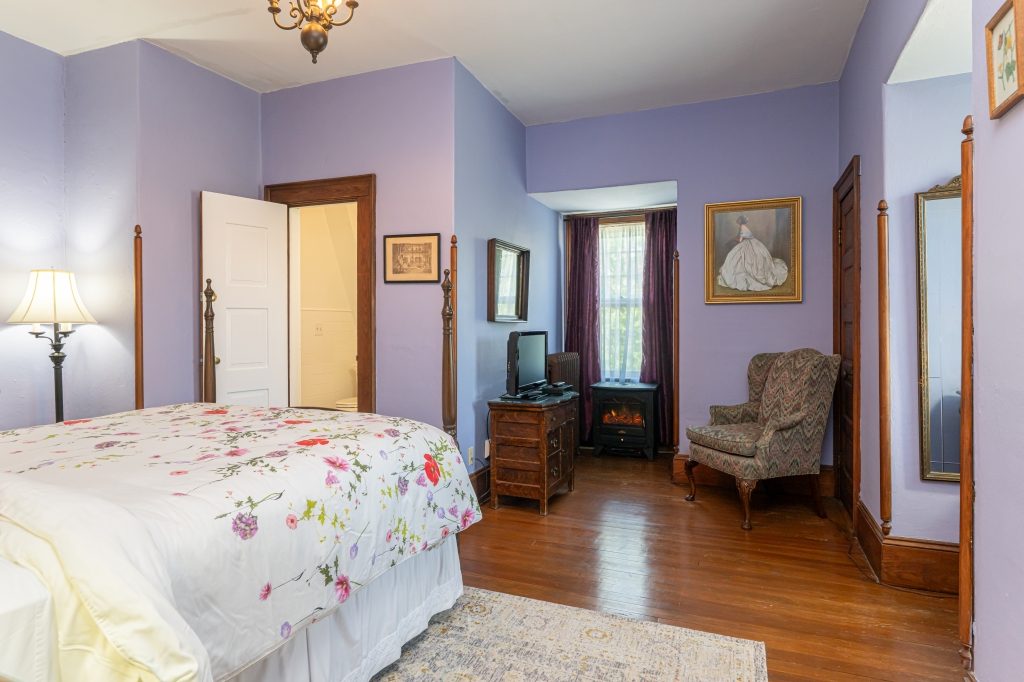 Maggie room — third-floor queen bed with floral bedspread, lavender walls, electric fireplace stove, and lady portrait at The Reynolds Mansion