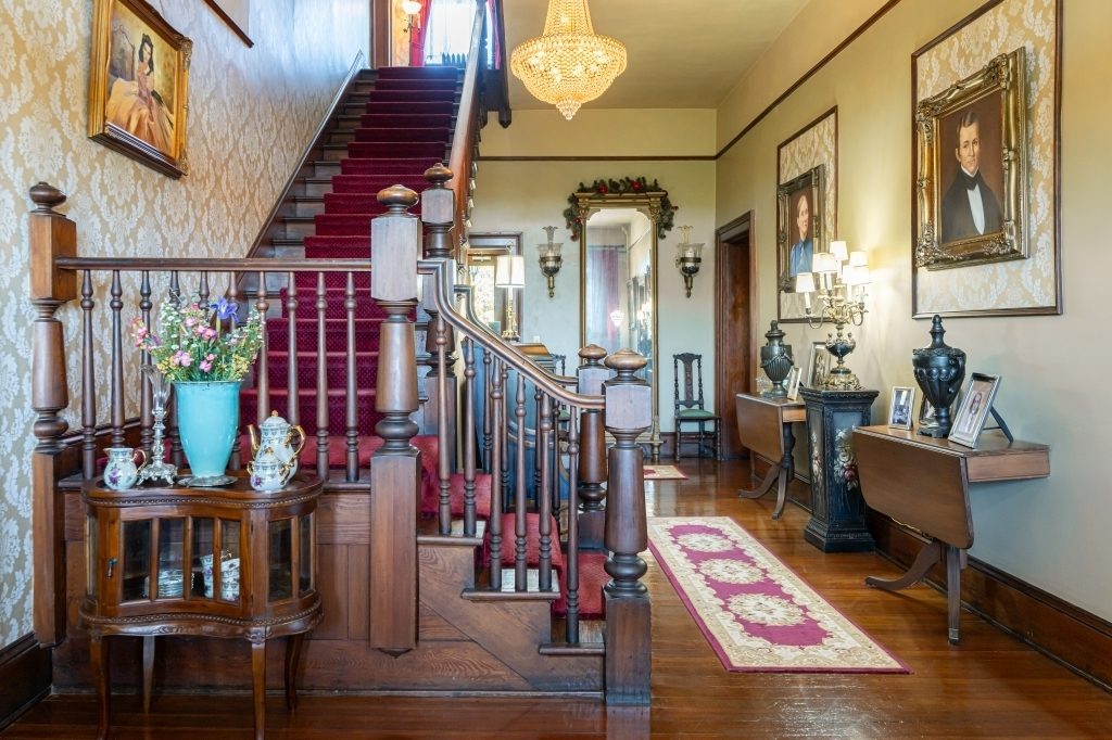 The grand staircase at The Reynolds Mansion — Colonial Revival staircase with turned balusters and crimson carpet runner, crystal chandelier, and period portraits flanking the wall in the entrance hall