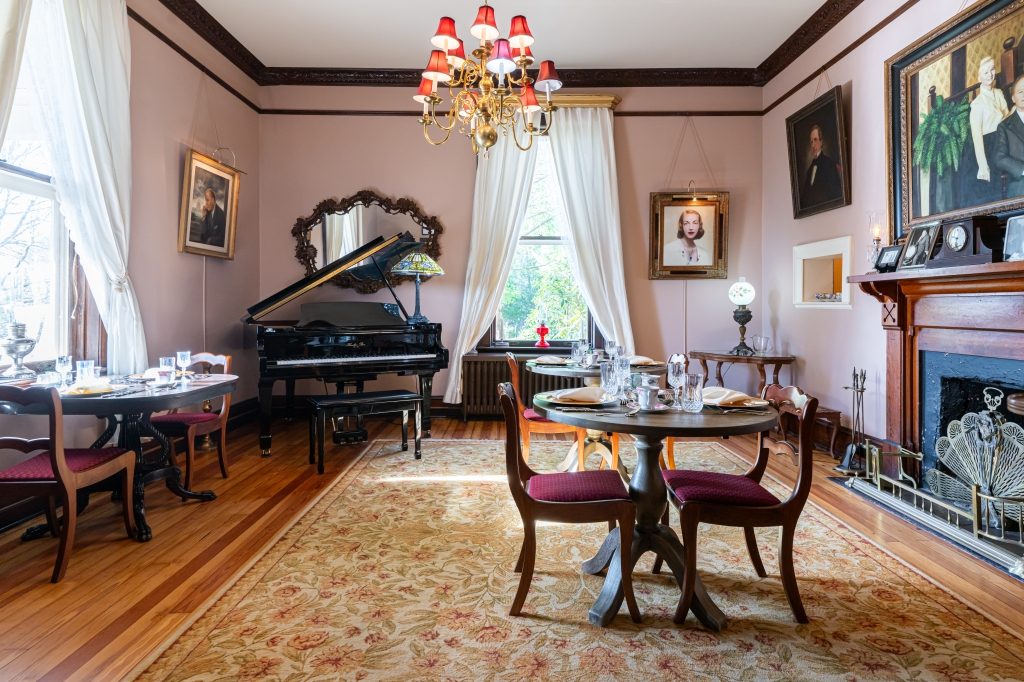 The parlor and dining room at The Reynolds Mansion — pink walls, period chandelier, grand piano, family portraits, and dining tables set for breakfast in the room Nathaniel Reynolds expanded around 1905