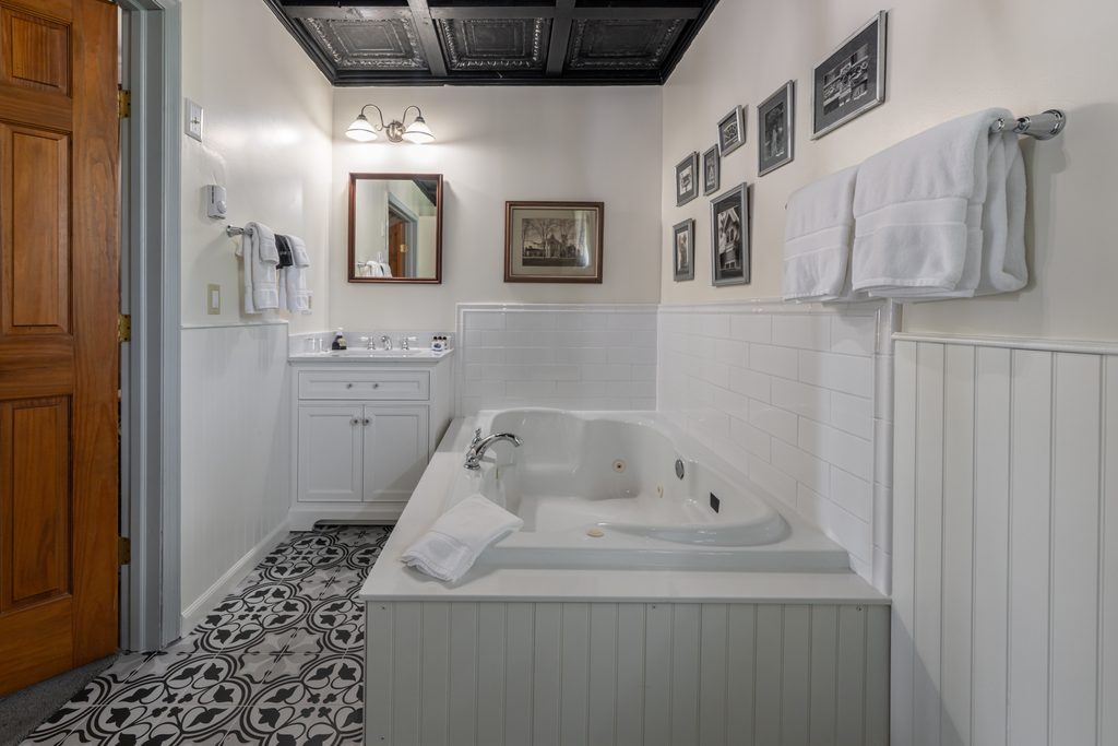 All That Jazz suite — white vanity with framed mirror beside the Jacuzzi tub, pressed-tin ceiling and patterned tile floor at The Reynolds Mansion