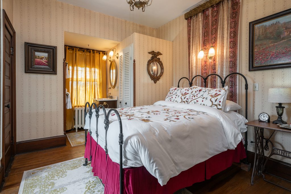 Inez room — iron bed with floral duvet and red bedskirt beside a curtained window with mountain light at The Reynolds Mansion