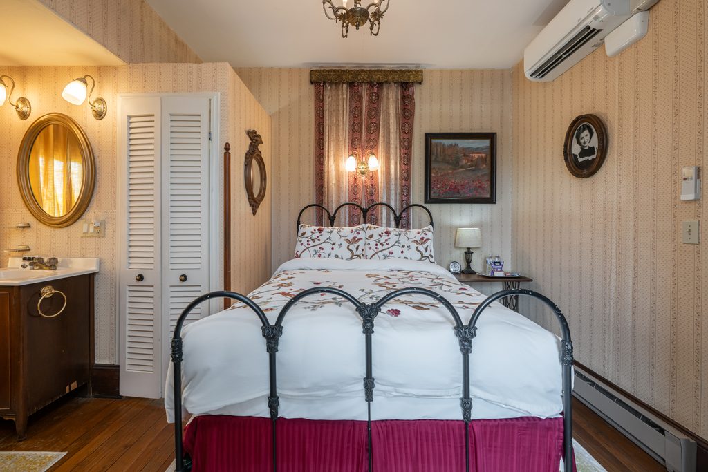 Inez room — antique iron bed with floral linens under a brass chandelier, framed landscape art on striped wallpaper at The Reynolds Mansion