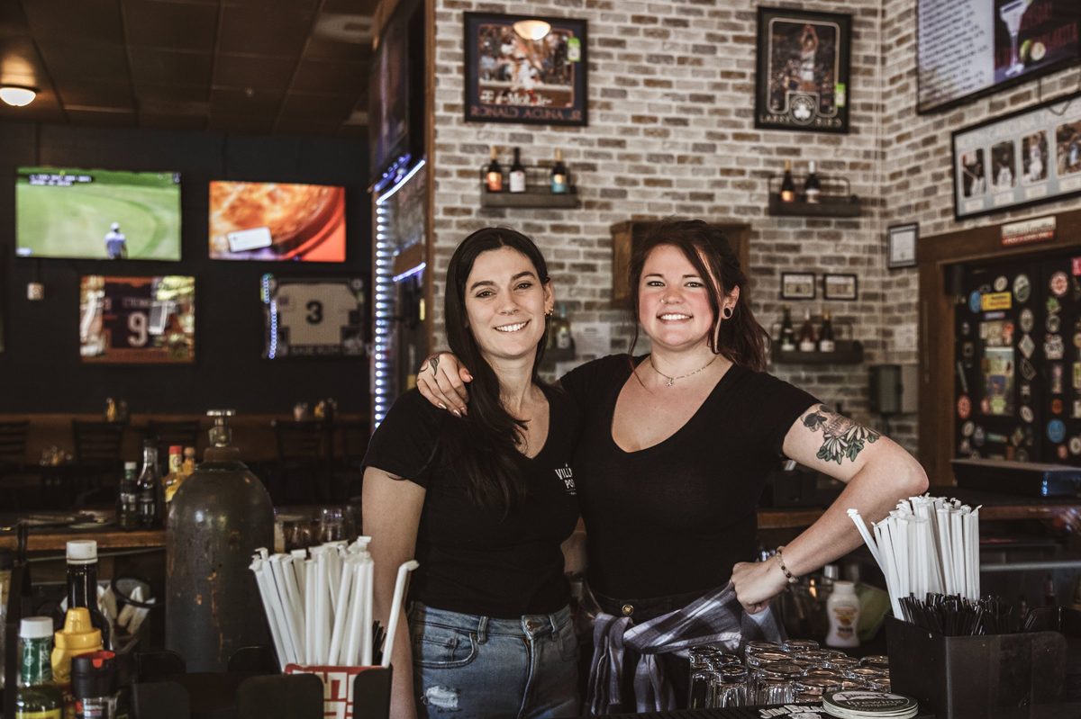 Village Porch bartenders behind the bar — TVs showing sports, brick walls, and a welcoming neighborhood pub atmosphere