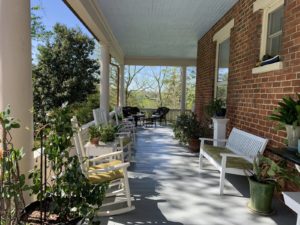 Sunny porch with white rocking chairs, brick walls, and a view of trees.