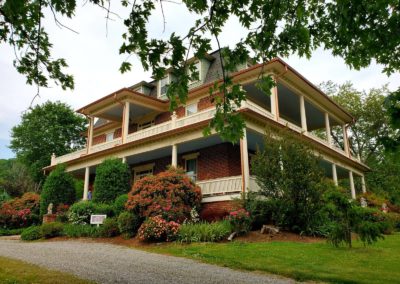 House with a large front porch and green lawn surrounded by trees.