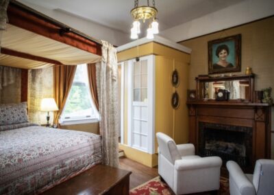 Elegant bedroom with bed, armchair, fireplace, and portrait above mantel, lit by a chandelier.