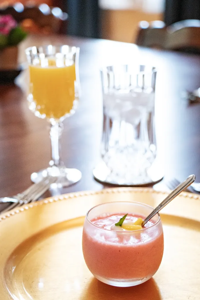 Pink smoothie in a glass with a spoon on a gold tray, with an out-of-focus orange juice glass behind.