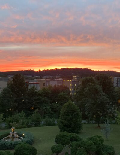 Sunset over a park with fountain and silhouette of buildings against a red-orange sky.