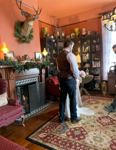 Man in vest standing in a vintage room with a fireplace, decorative mantle, and a person in the background.